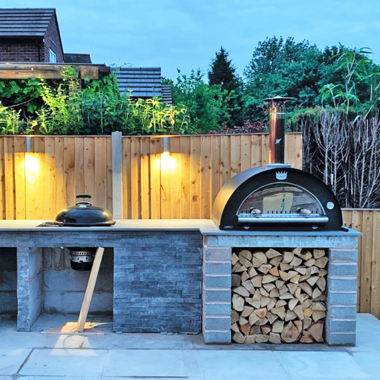 Clementi Wood Fired Pizza Oven 100x80 shown situated in garden on shelf above logs. The Outdoor Grill Company.