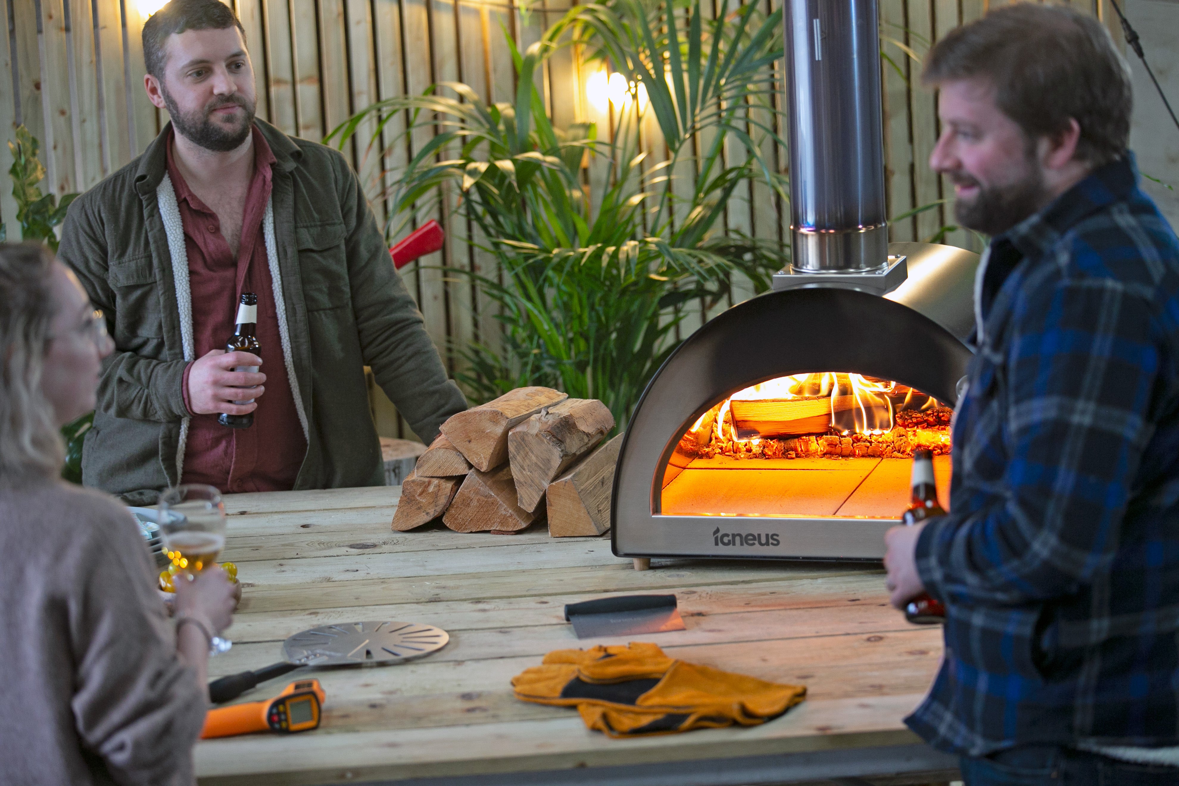 two men and a lady chatting with a glass of wine standing around the Igneus Classico which is fired up ready to add the food